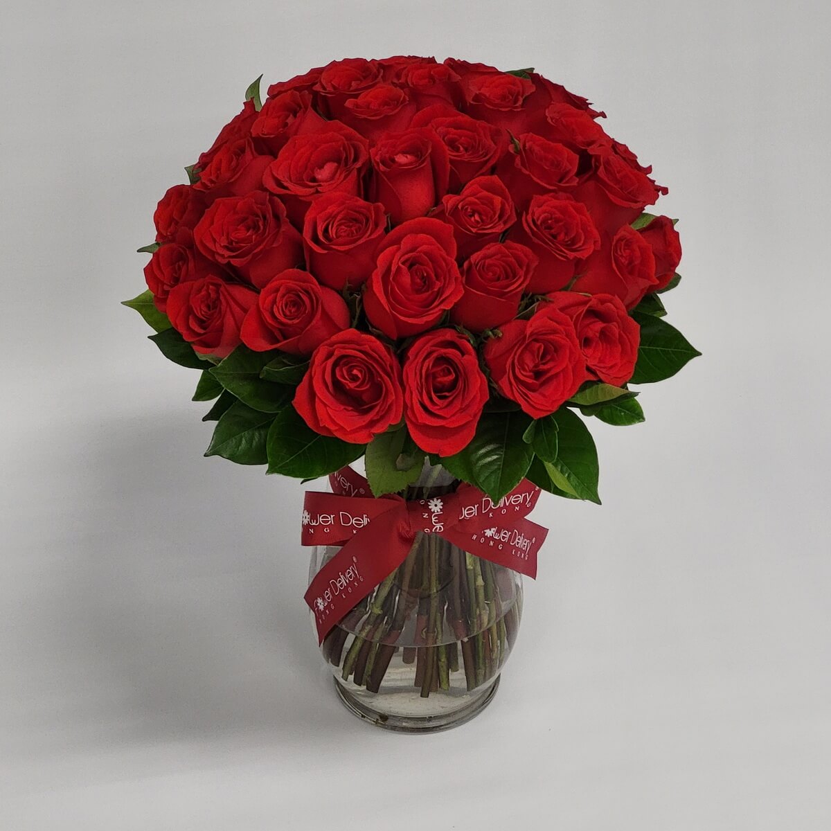 Classic bouquet of deep red roses arranged in a rounded dome shape, accented with glossy greenery and placed in a clear glass vase with a red ribbon. 經典紅玫瑰圓形花束，搭配翠綠葉片，置於透明玻璃瓶中，繫上紅色緞帶，象徵熱情與浪漫。