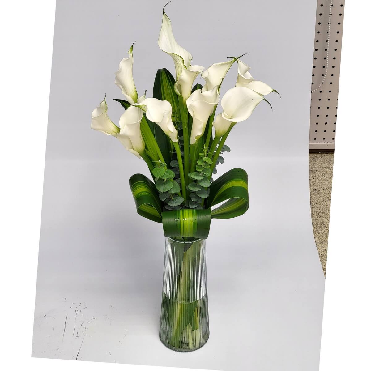 Elegant arrangement of white calla lilies with eucalyptus and wide looped ti leaves in a tall ribbed glass vase. 純白馬蹄蓮搭配尤加利葉與環繞的翠綠葉片，插於高透明條紋玻璃瓶中，展現高雅與現代感。