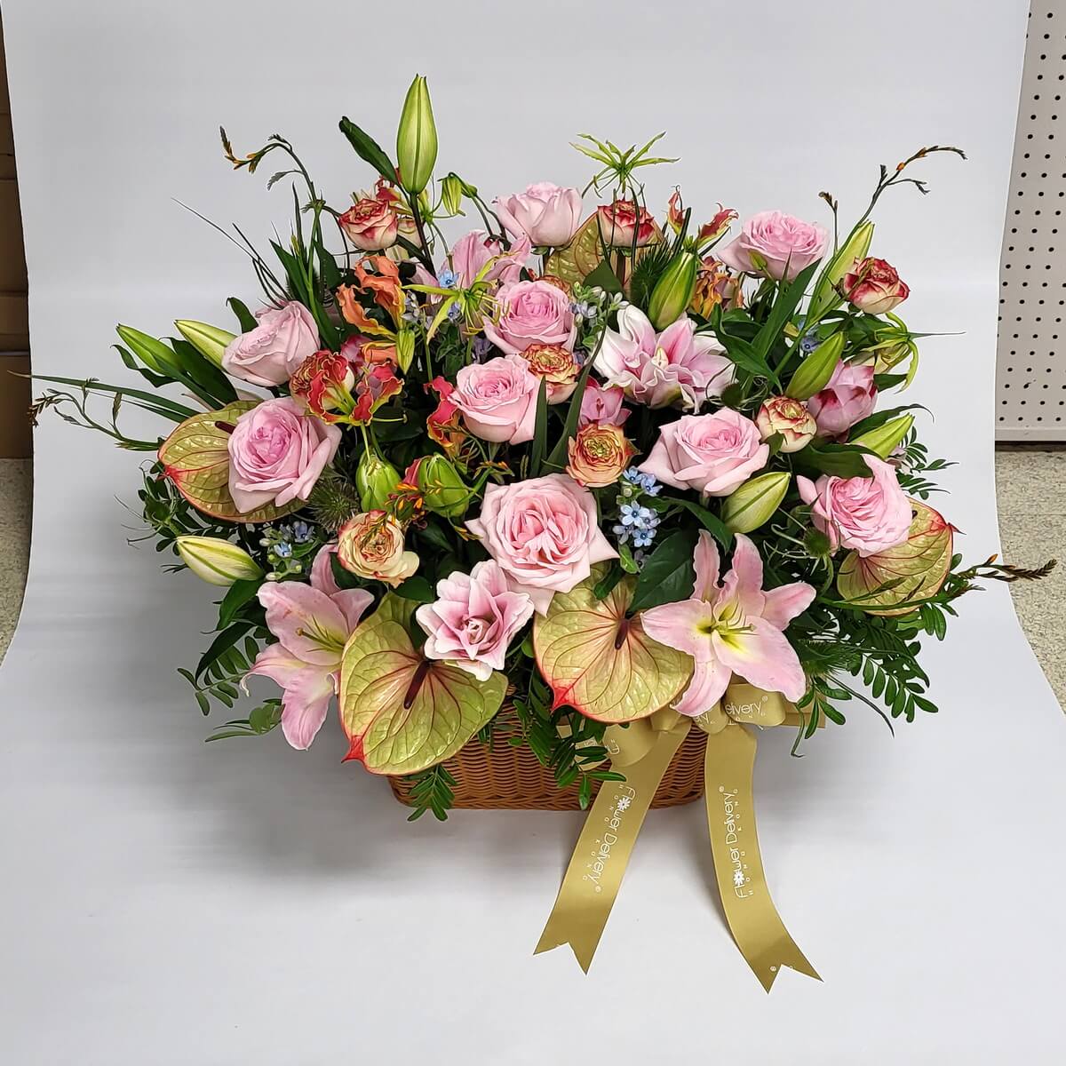 Pink roses, lilies & champagne anthuriums in basket 粉紅玫瑰與百合花為主角，配以香檳火鶴花、康乃馨與藍色小花，綠意點綴，花籃金緞帶，設計溫柔浪漫。