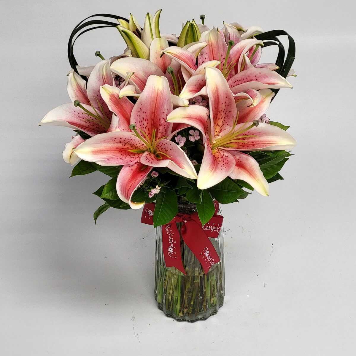 Pink stargazer lilies with waxflowers 粉紅星百合盛開，搭配蠟梅與綠葉，展現華麗優雅的風格，置於雕紋玻璃瓶中，紅色緞帶增添喜慶氛圍。