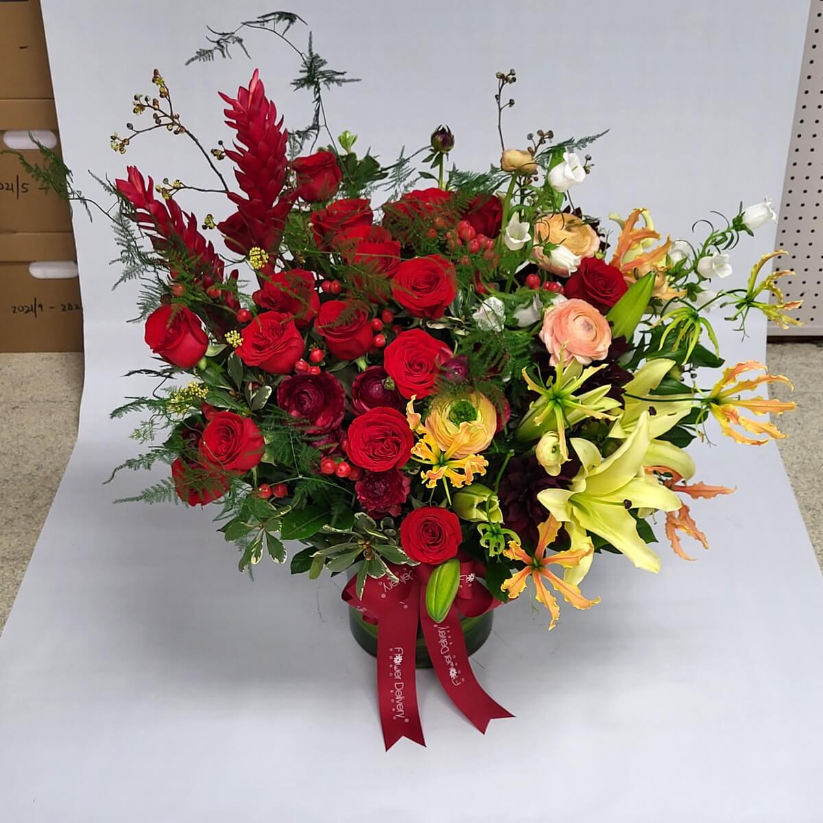 Vibrant floral mix with red roses, yellow lilies, ranunculus & tropical accents 豐盛紅玫瑰、百合、金魚草與熱帶花卉搭配，插於圓瓶中，紅絲帶點綴，氣勢非凡。