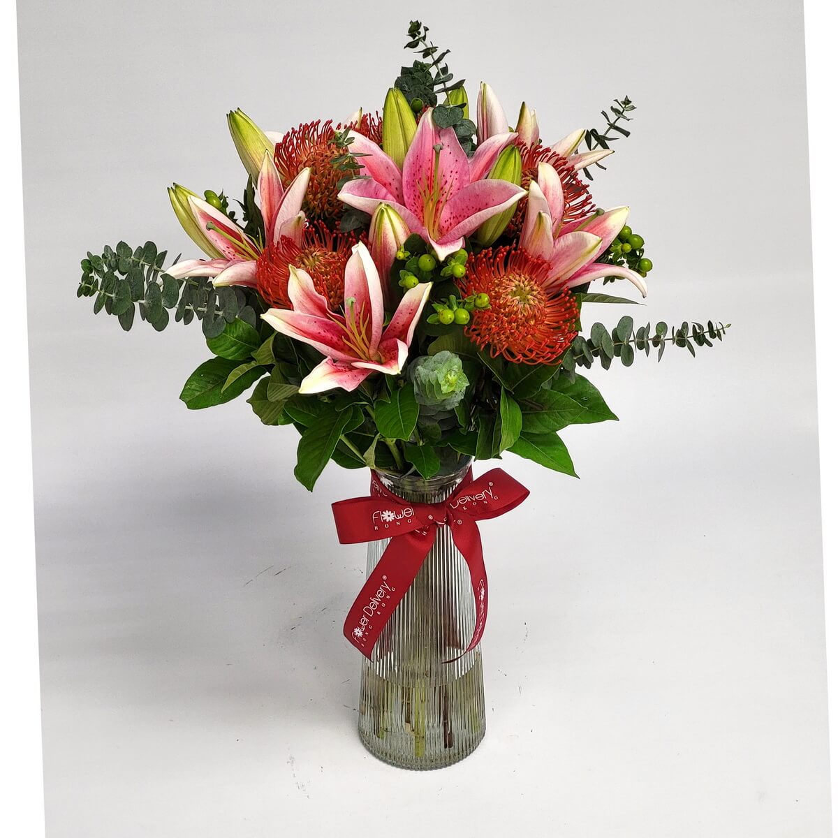 Bold bouquet of pink lilies, orange pincushion proteas, and lush greens in a tall vase 熱情粉百合與針墊花搭配綠葉，插於高身條紋玻璃瓶中，繫上紅色蝴蝶結，展現鮮明活力。