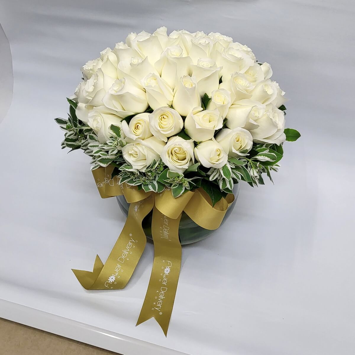 Classic white rose dome arrangement 白玫瑰圓頂花藝，層層堆疊展現豐盈質感，點綴斑葉綠植，圓形玻璃花瓶配金色緞帶，展現優雅純潔之美。