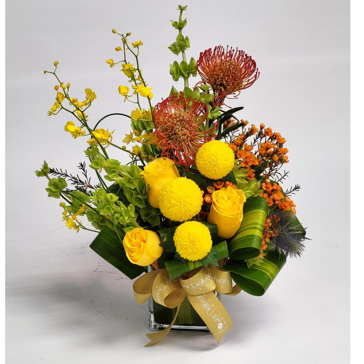 Full flower arrangement in glass vase featuring yellow button chrysanthemums, yellow roses, orange pincushion protea, orange waxflowers, yellow orchids and green bells of Ireland - 玻璃瓶插花包括黃色乒乓菊、玫瑰、橙色針墊花、蠟花、黃色蘭花與愛爾蘭鈴
