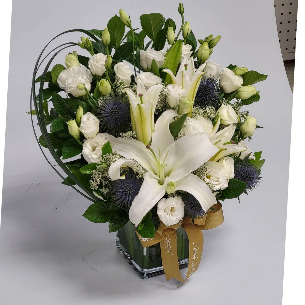Elegant white lilies and roses 百合與白玫瑰綻放間，紫色刺球花增添質感，綠葉點綴，搭配金色絲帶與玻璃瓶，展現純淨與尊貴的和諧花藝。