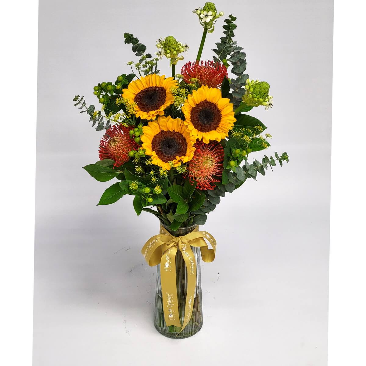 Sunflower and protea arrangement 向日葵搭配紅色針墊花、尤加利葉與多種綠植，高身玻璃瓶綁金色絲帶，展現熱情明亮的夏日氛圍，適合送給充滿活力的朋友。