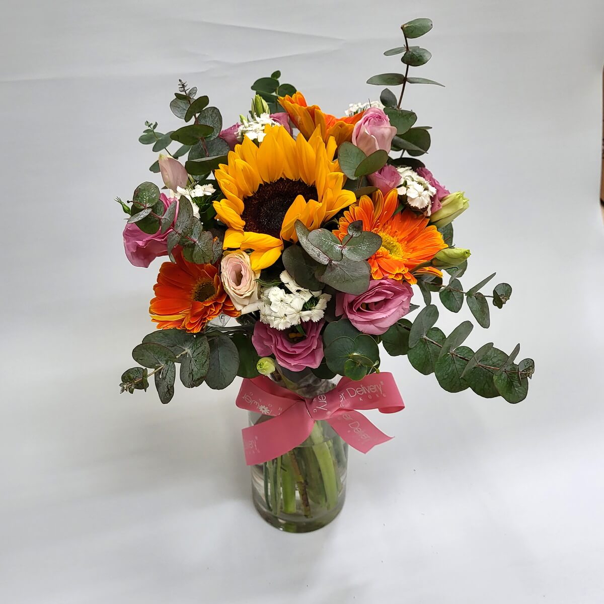 Sunflowers, gerberas, roses with eucalyptus 向日葵與橙色太陽花、粉玫瑰及白色配花交織，搭配尤加利葉，插於玻璃瓶中，綁上粉紅絲帶，散發活力與浪漫氣息。