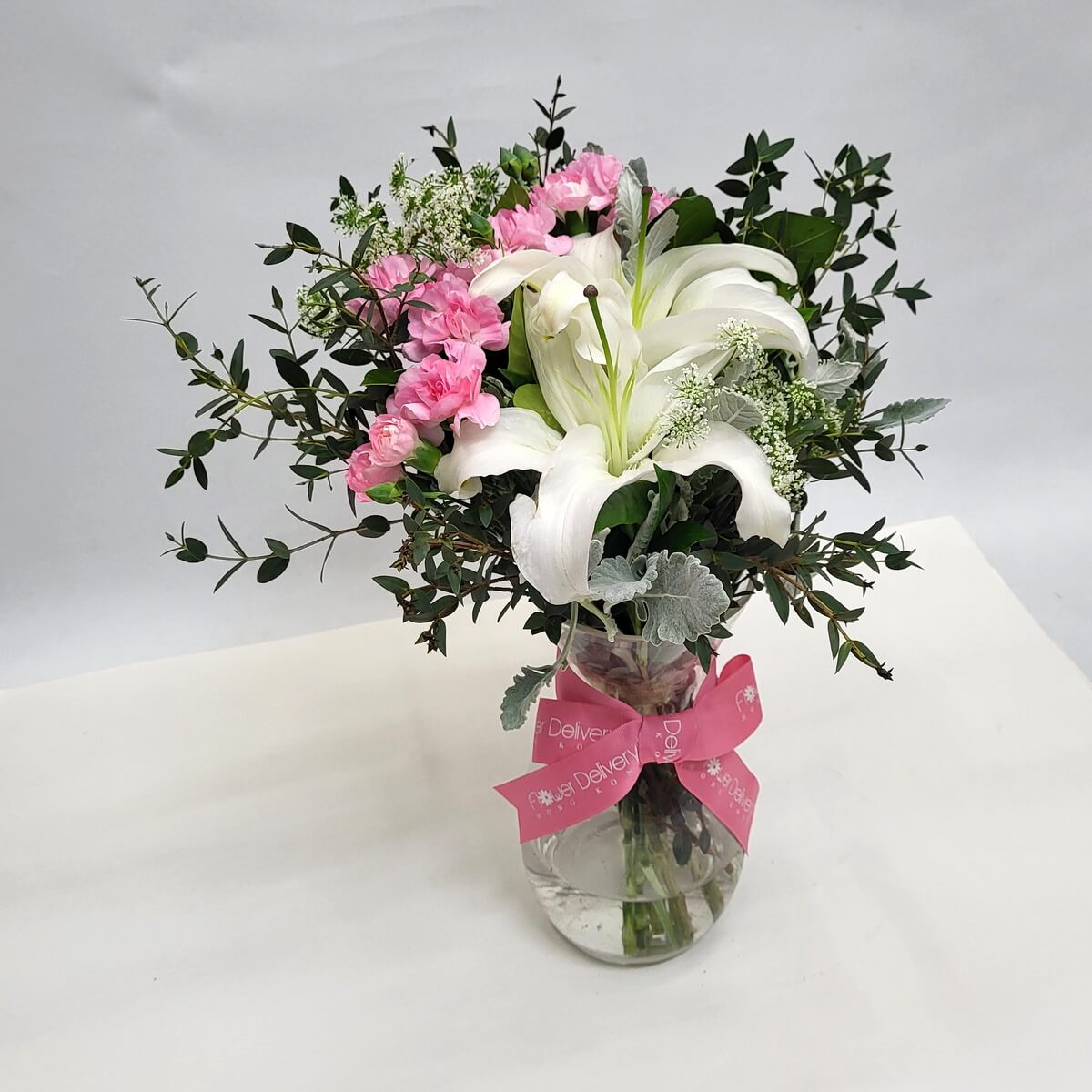 Elegant mix of white lilies and pink carnations with assorted foliage in a glass vase 白百合與粉康乃馨點綴多種綠葉，裝於玻璃瓶中，粉紅絲帶增添溫柔氣息。