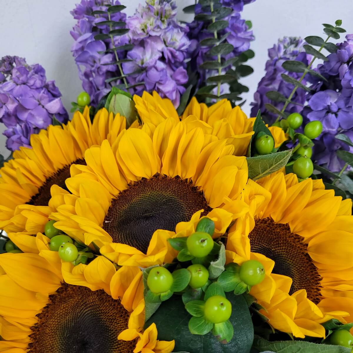 Zoomed-in arrangement showing yellow sunflowers, soft purple stock flowers, and clusters of green hypericum berries 向日葵、柔紫松葉牡丹及成簇綠色貝母果的近距離插花構圖