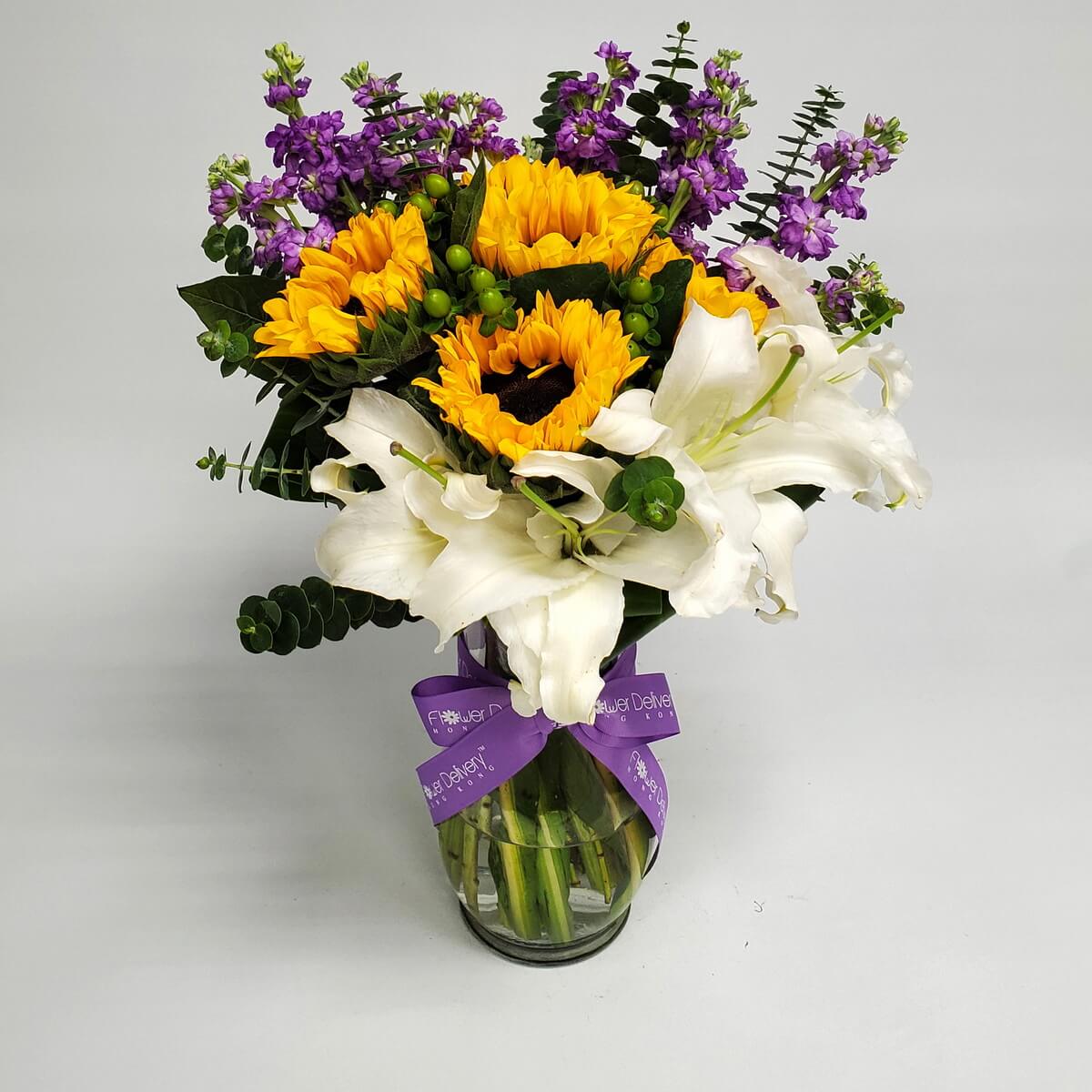 Sunflowers, lilies, and purple flowers elegantly arranged in a vase with purple ribbon 向日葵搭配百合與紫花，擺放於透明花瓶中，紫色絲帶點綴，展現優雅氣息。