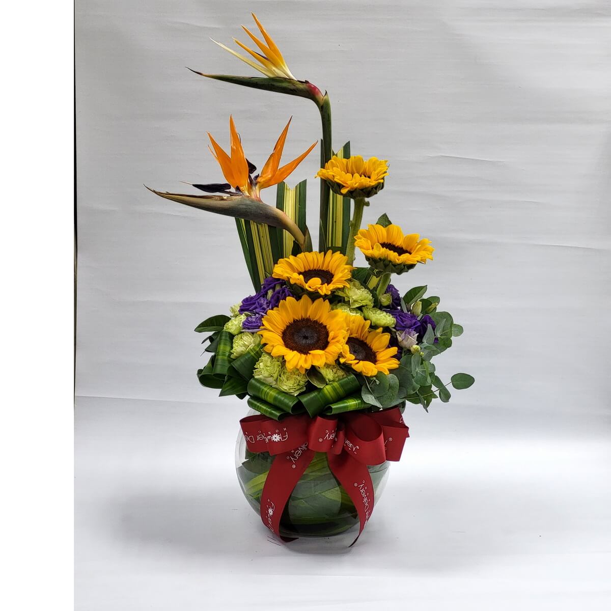 Striking sunflowers and birds of paradise in a globe vase with purple and green accents, styled with tropical leaves and red ribbon 向日葵與天堂鳥花藝，搭配紫花與青綠葉材，置於圓形玻璃花瓶中，紅色緞帶增添喜慶氣氛。