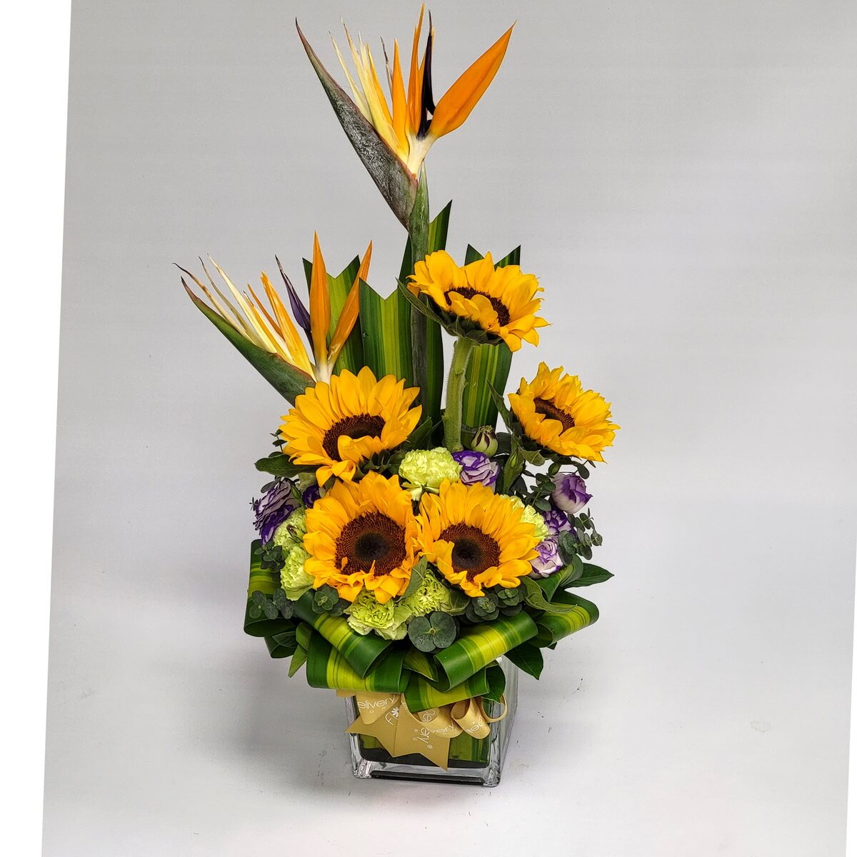 Vertical floral design featuring sunflowers, bird of paradise, green carnations, purple-white lisianthus, and tropical greens in a glass vase 向日葵、天堂鳥、綠康乃馨、紫白洋桔梗與熱帶綠葉構成的玻璃瓶直線花藝設計