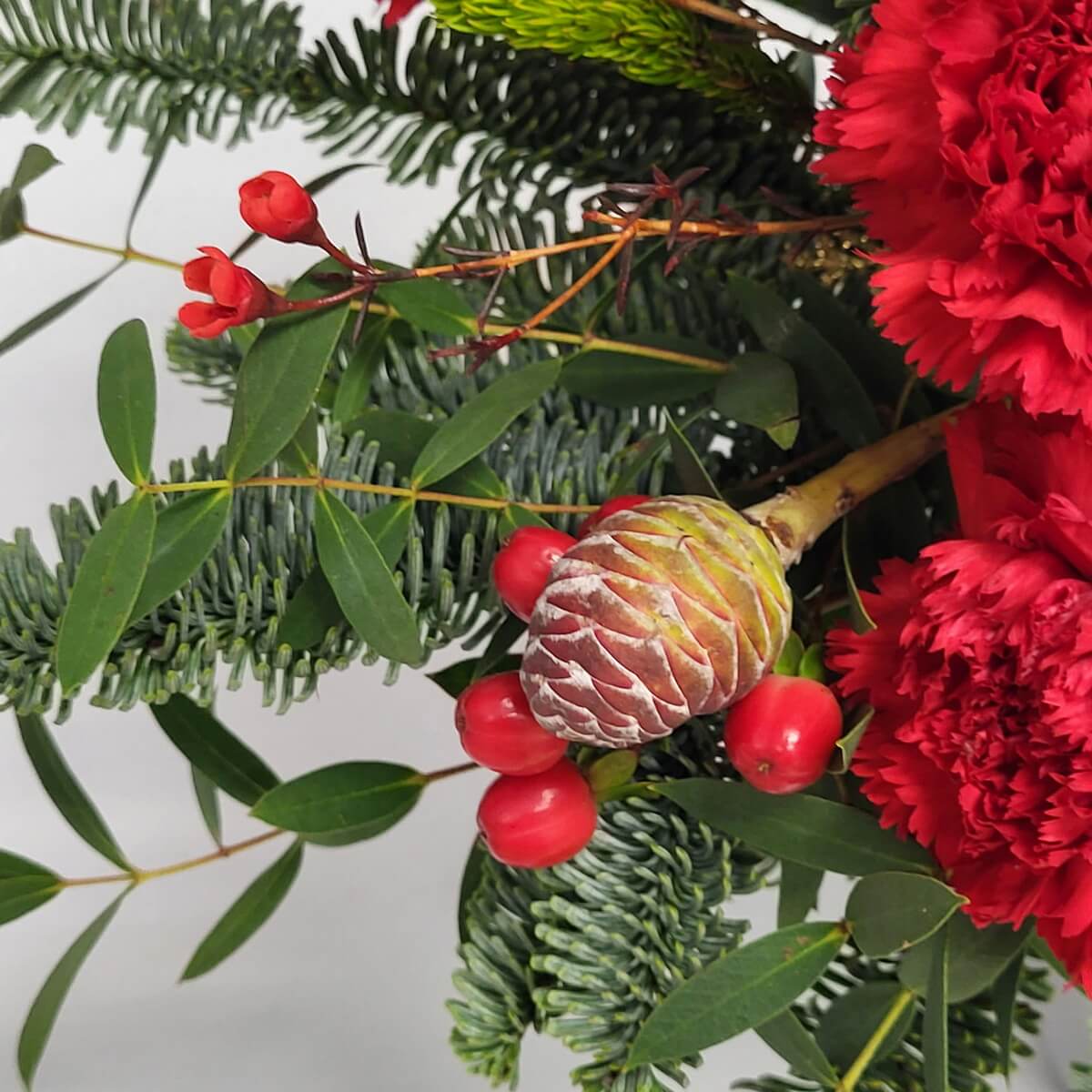 Close-up showing red carnations, pine cone, red hypericum berries, red waxflowers, and fir greenery in a festive floral mix - 紅康乃馨、松果、紅繡球果、紅蠟花及冷杉葉的花藝特寫