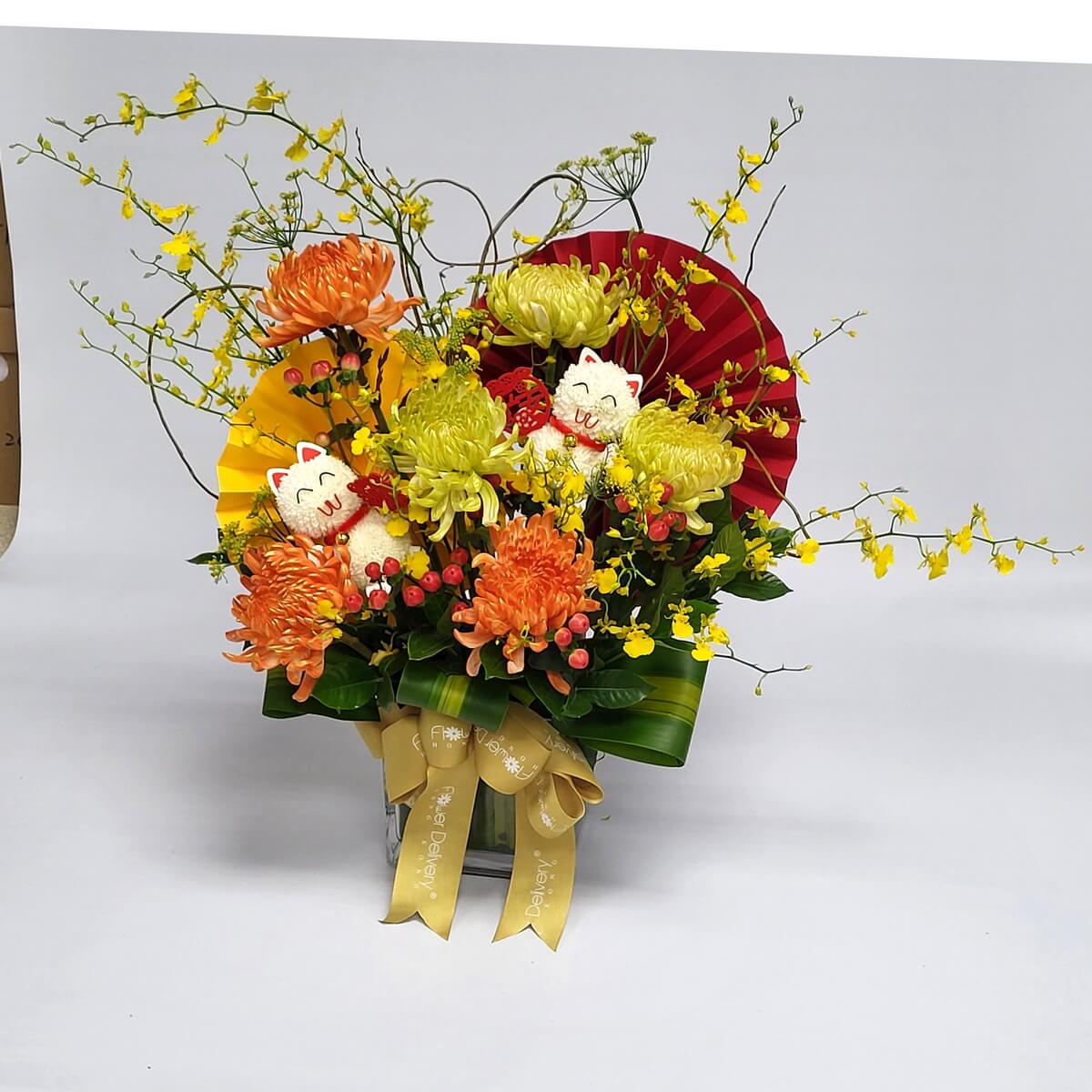 Bright chrysanthemum mix with oncidium orchids, red berries, fan-shaped paper backdrops, and lucky cat picks in a square vase 橙黃菊花、舞春蘭、紅漿果搭配紅黃紙扇與可愛招財貓擺件，方形玻璃花瓶設計，充滿節慶氣氛。