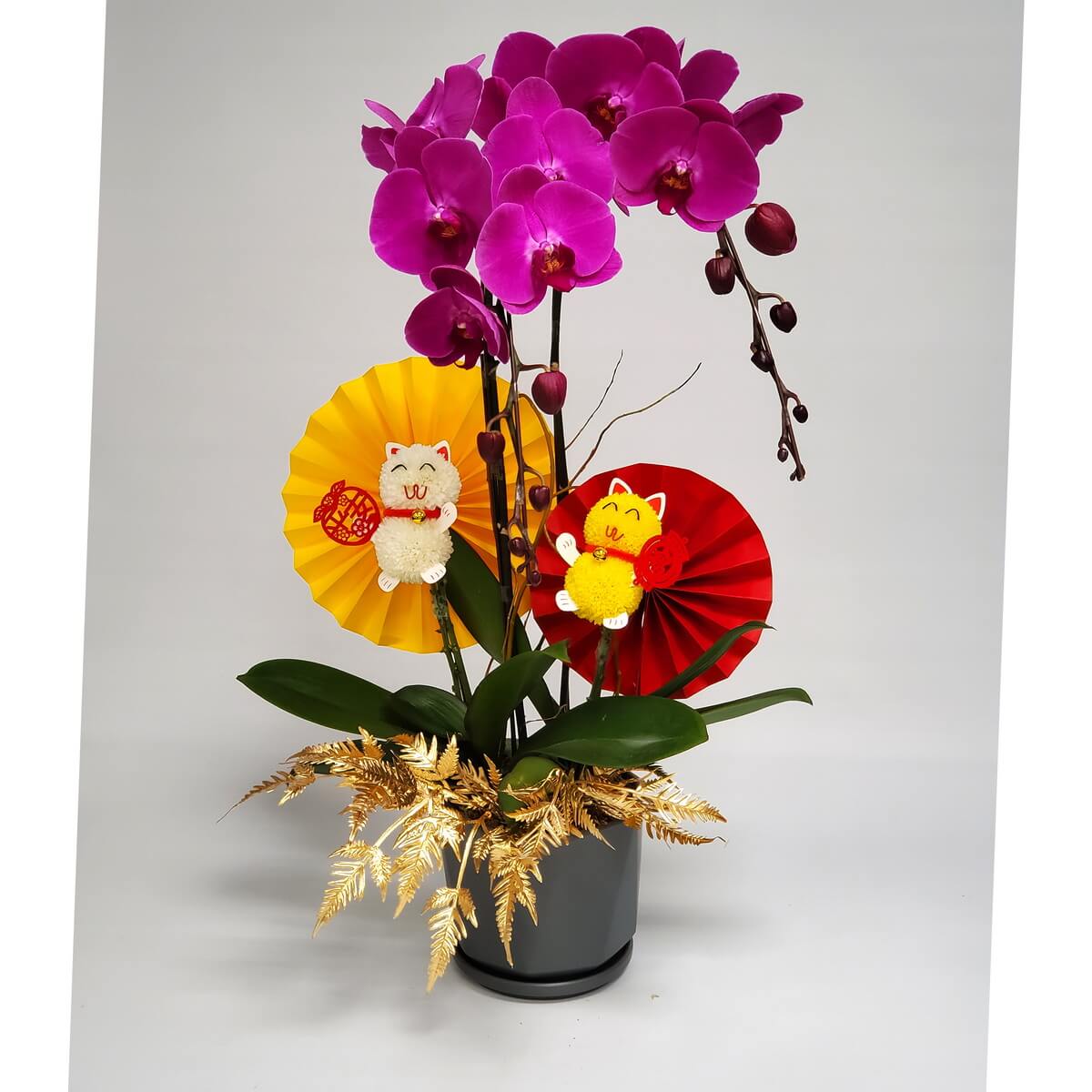 Arrangement of magenta Phalaenopsis orchids with hanging buds, gold foliage, festive paper fans, and cat decorations in grey pot 洋紅蝴蝶蘭盆栽，配垂掛花苞、金色蕨葉、紅黃節慶摺紙扇與招財貓裝飾，擺放於灰色花盆中。