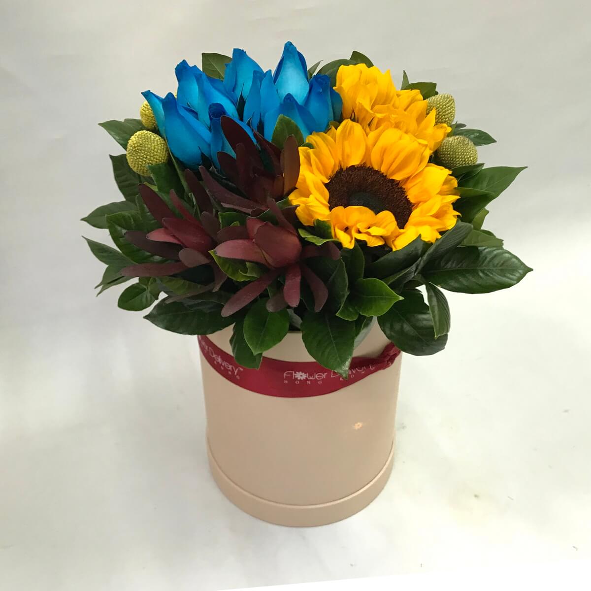 Hatbox arrangement with yellow sunflowers, blue roses, billy balls, burgundy accents, and green foliage 向日葵及藍玫瑰為主體，配搭黃金球花、紅褐葉材與亮綠葉片，擺設於米色圓形花盒中。