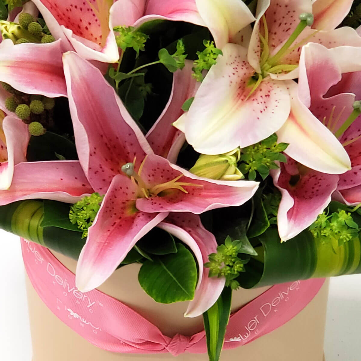 Detail view of blooming pink stargazer lilies with speckled petals, accented by green fillers and structured tropical foliage in a pink ribbon hatbox - 粉紅星形百合綻放，配斑點花瓣、綠色配花與造型熱帶葉，粉紅絲帶花盒裝飾