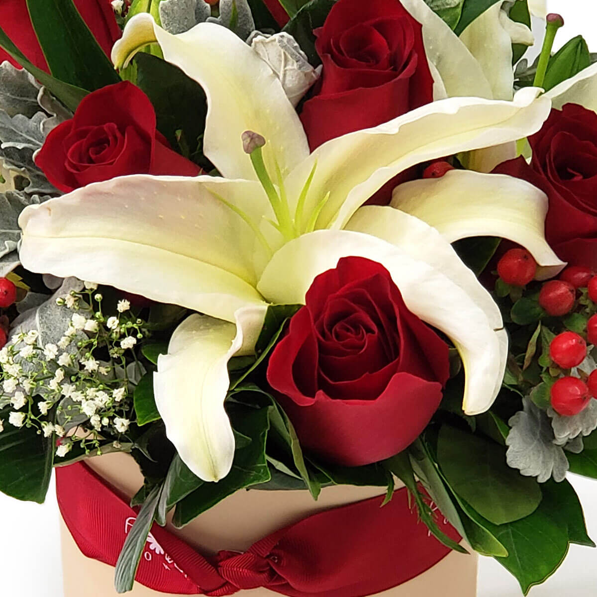 Detail view of an arrangement featuring white oriental lilies, red roses, baby's breath, red hypericum berries, and silver dusty miller in a round floral box - 白東方百合、紅玫瑰、滿天星與紅果搭配銀葉菊，圓形花盒設計的花藝細節圖