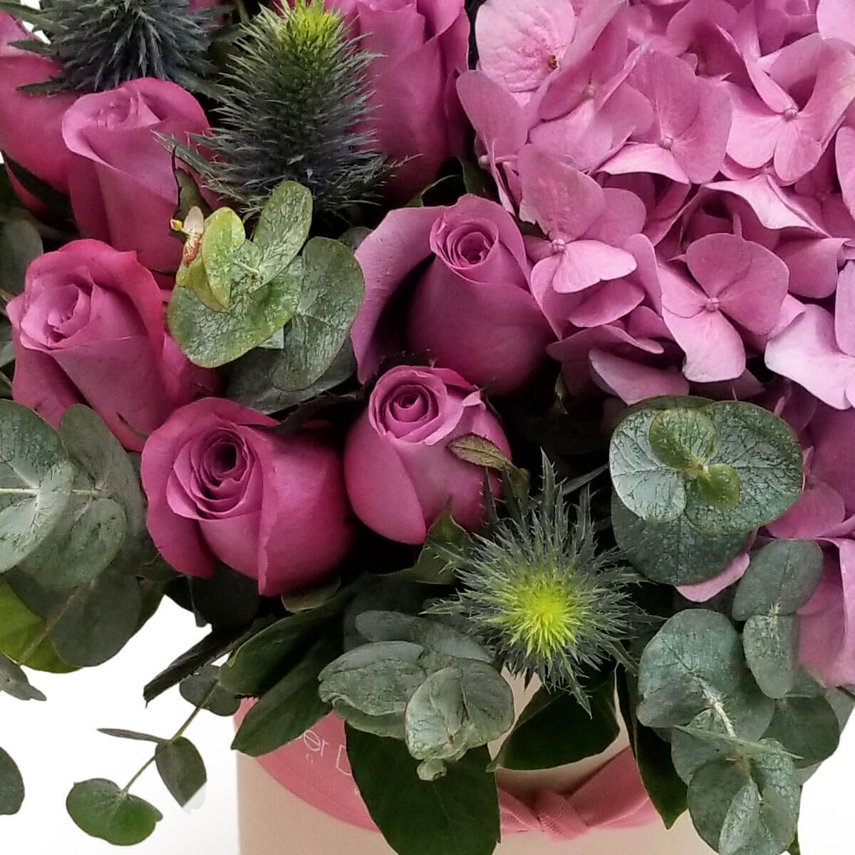 Detail view of a floral arrangement featuring mauve roses, soft pink hydrangeas, thistle-like eryngium, and silvery eucalyptus foliage in a blush-toned box - 粉紫玫瑰、柔粉繡球花、刺狀松葉牡丹與銀灰尤加利葉搭配，粉色花盒花藝細節圖