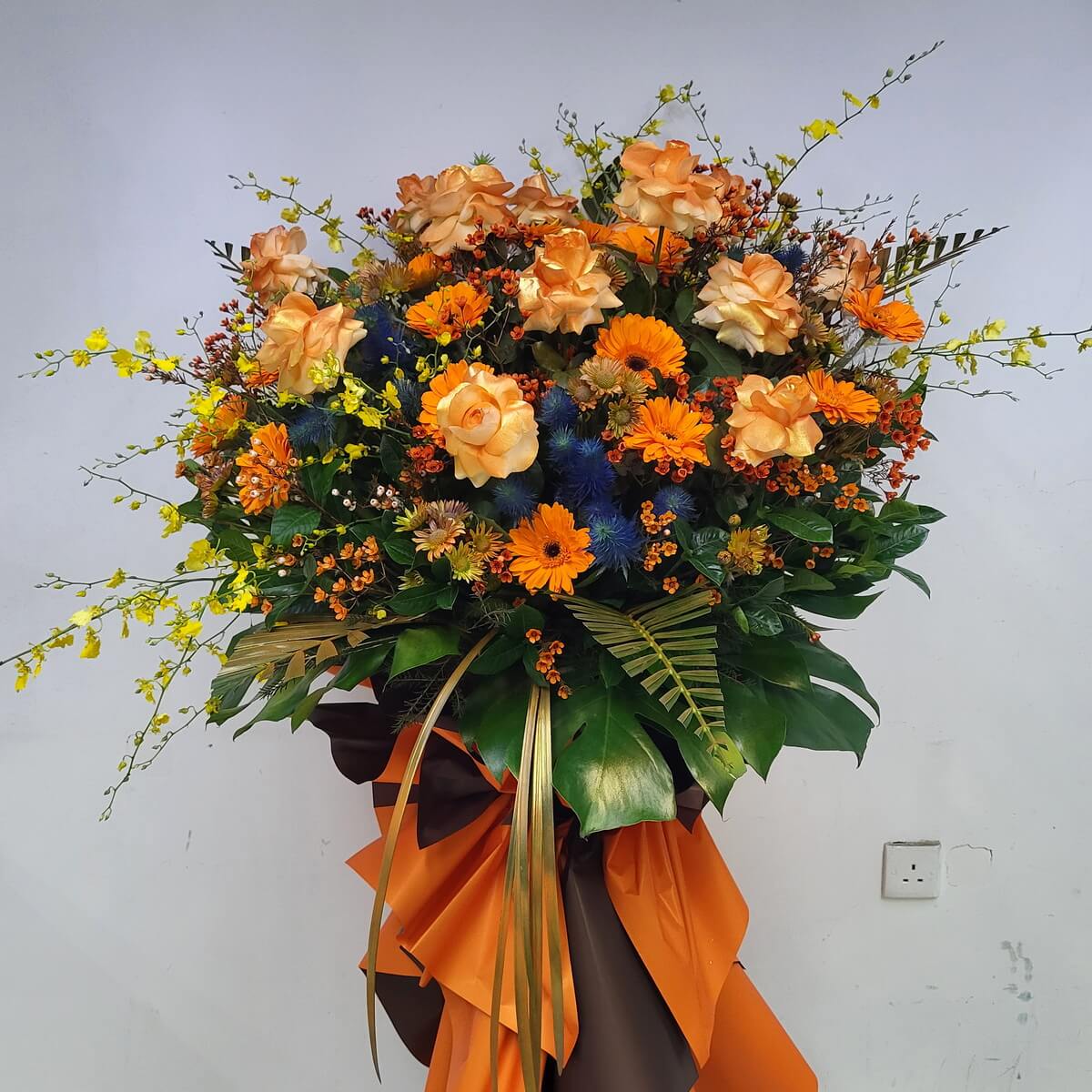 Full standing spray featuring orange roses, bright orange gerberas, yellow oncidium, blue thistles, waxflowers, and palm leaves with orange-brown wrapping - 橙玫瑰太陽菊黃文心蘭藍薊蠟花與棕櫚葉配橙啡包裝企牌花籃