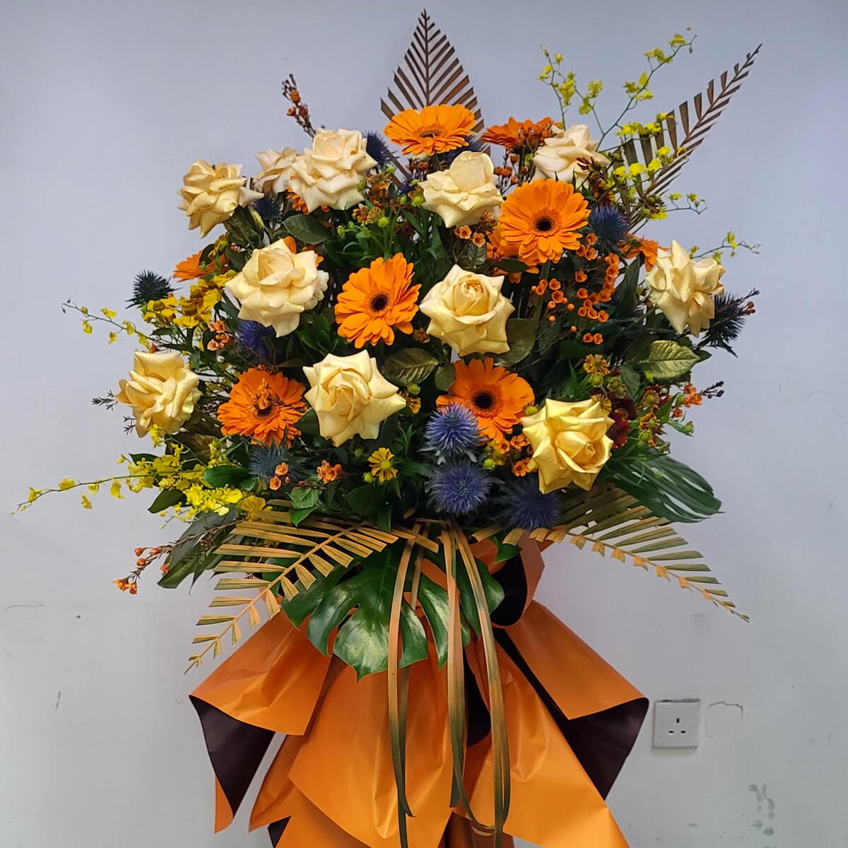 A vibrant floral stand featuring orange gerberas, champagne roses, yellow oncidium orchids, blue thistles, and golden foliage.
橙香檳玫瑰、跳舞蘭與藍色薊花組成活力滿滿的花牌，金葉點綴，襯以大橙蝴蝶結，展現熱烈氣氛。