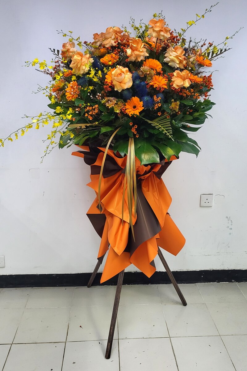 Complete standing floral display with orange roses, bright gerberas, yellow oncidium orchids, blue thistles, waxflowers, and tropical foliage, wrapped in orange and brown - 橙玫瑰太陽菊黃文心蘭藍薊蠟花熱帶葉材配橙啡色包裝企牌花籃