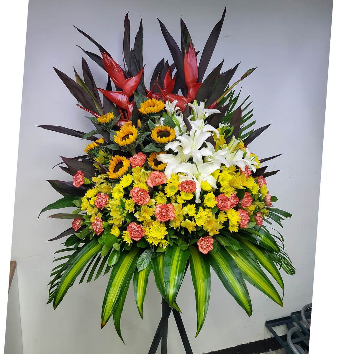 A bold floral stand featuring yellow sunflowers, white lilies, orange carnations, yellow button chrysanthemums, and red heliconia with tropical foliage.
配以向日葵、百合、橙康乃馨、黃色乒乓菊與天堂鳥，氣勢澎湃，綠葉扶襯。