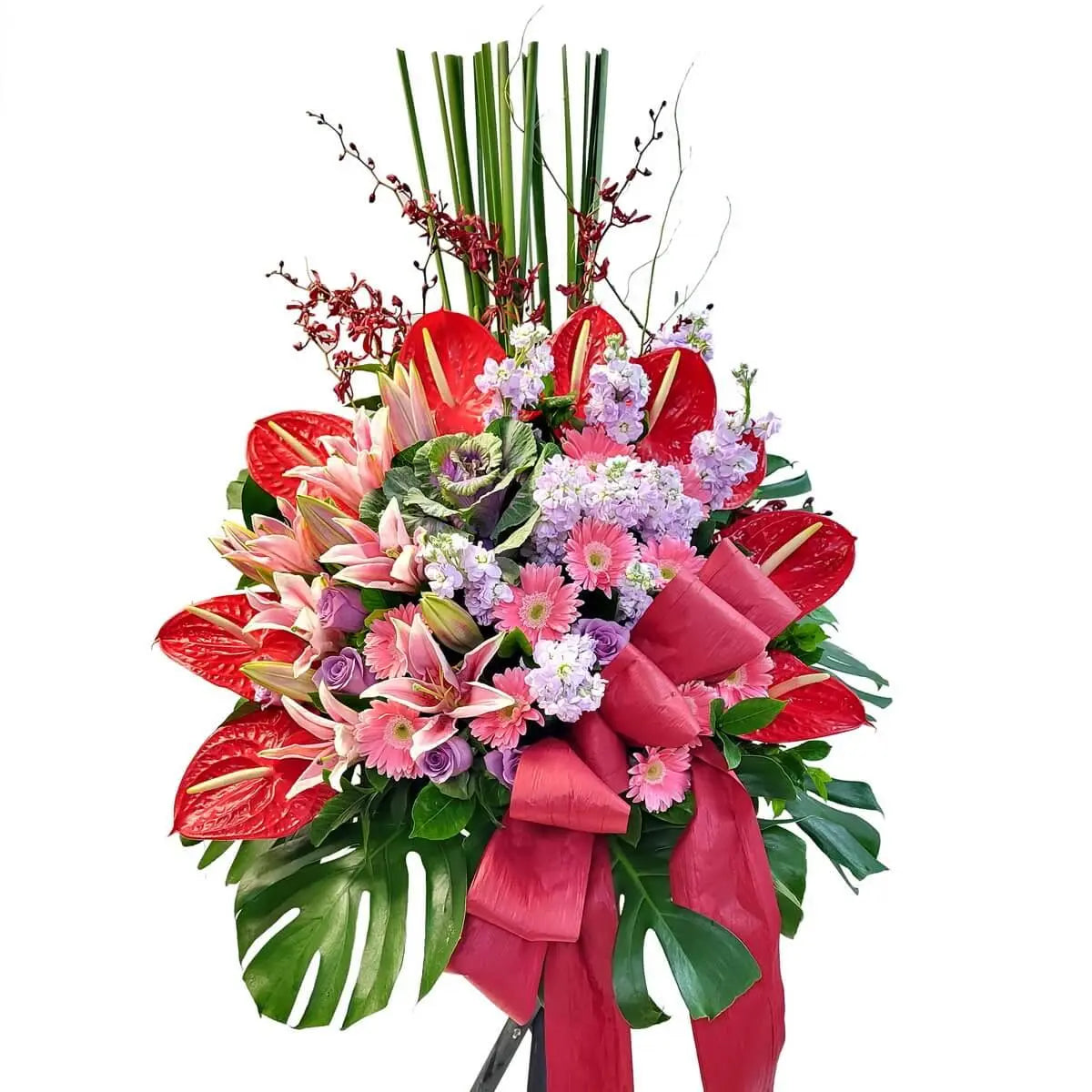 Standing arrangement with red anthuriums, pink lilies, gerberas, purple roses, light pink stock flowers, and mixed foliage - 花牌設計含紅火鶴、粉百合、粉非洲菊、紫玫瑰及粉紫紫羅蘭，配以熱帶葉材與大紅蝴蝶結裝飾。