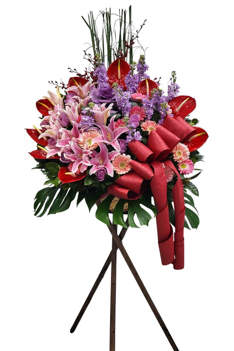 Standing floral arrangement featuring pink lilies, purple lisianthus, red anthuriums, pink gerberas, and lavender stock, accented with monstera leaves and large red ribbon - 粉百合、紫洋桔梗、紅掌、粉扶郎與紫花鬱金香花牌，襯以龜背葉與大型紅絲帶