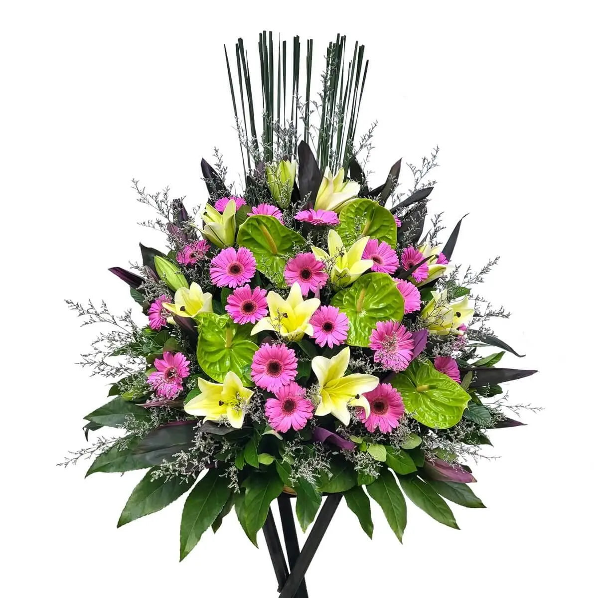 Standing floral display featuring yellow lilies, pink gerberas, green anthuriums, and layered greenery on a black tripod stand - 花牌設計配黃百合、粉紅非洲菊、綠火鶴與多款葉材，層次豐富，擺放於黑色三腳支架上。