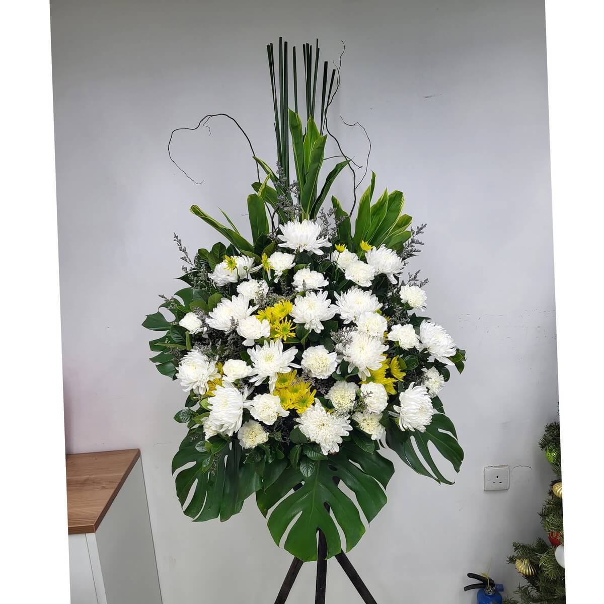 A solemn condolence stand of white and yellow chrysanthemums, enhanced with layered tropical greens and upright foliage. 白黃菊花搭配挺立綠葉與層次枝葉，呈現莊重悼念之意。