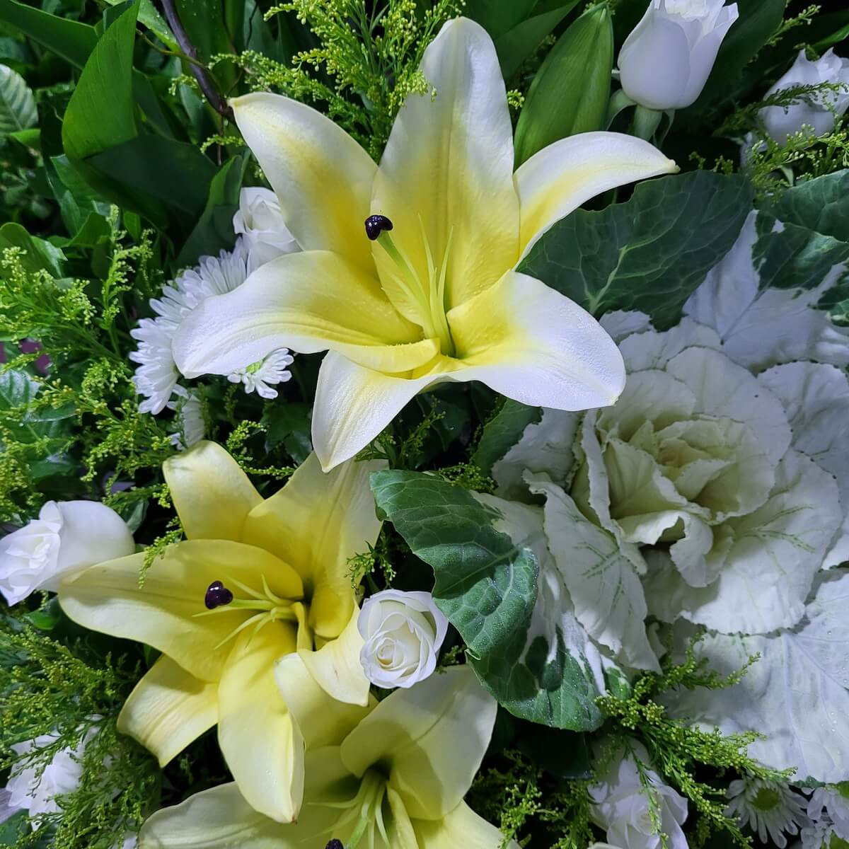Floral arrangement with yellow lilies, white roses, white chrysanthemums, ornamental cabbage, and assorted greens. | 黃百合、白玫瑰、白菊、羽衣甘藍及多款綠葉花藝設計。
