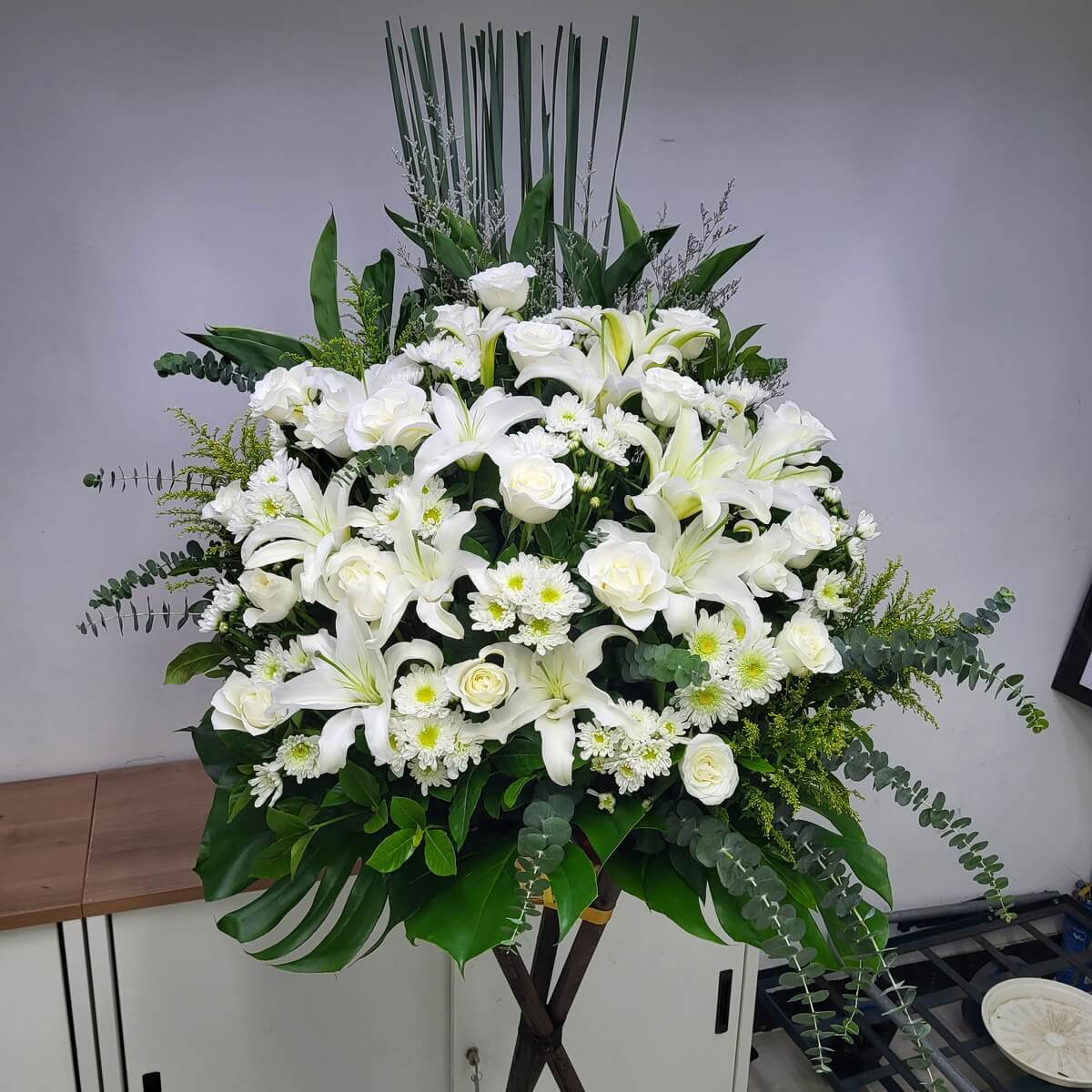 Formal standing arrangement featuring white lilies, roses, and button chrysanthemums, complemented by eucalyptus and tropical foliage 白百合、白玫瑰與白乒乓菊主體花材，搭配尤加利與熱帶葉材的正式直立花牌
