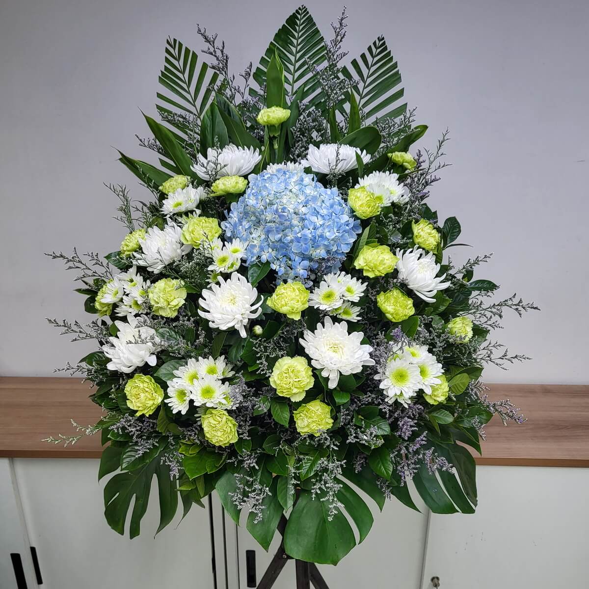 Layered standing arrangement featuring blue hydrangea, white spider and button chrysanthemums, green carnations, and tropical foliage 藍繡球、白蜘蛛菊、白乒乓菊、綠康乃馨與熱帶葉材構成的分層式直立花牌設計