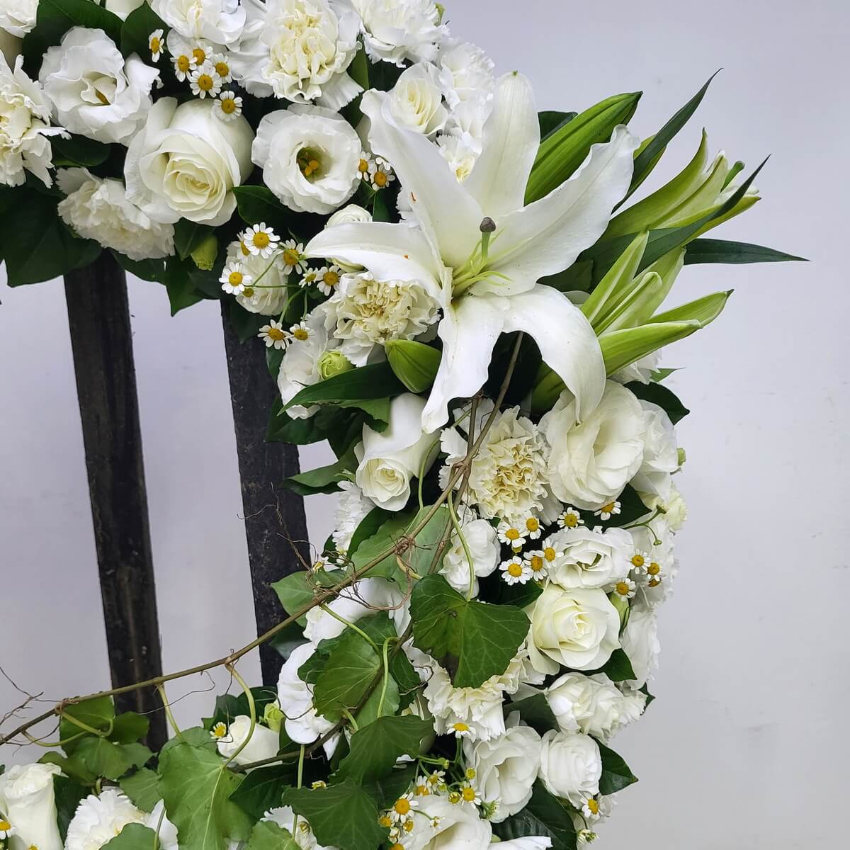 Close-up view of wreath featuring white lilies, roses, lisianthus, carnations, small chamomile flowers with yellow centers, and trailing ivy - 白百合、玫瑰、洋桔梗、康乃馨、黃芯小雛菊配垂藤裝飾花圈局部特寫畫面