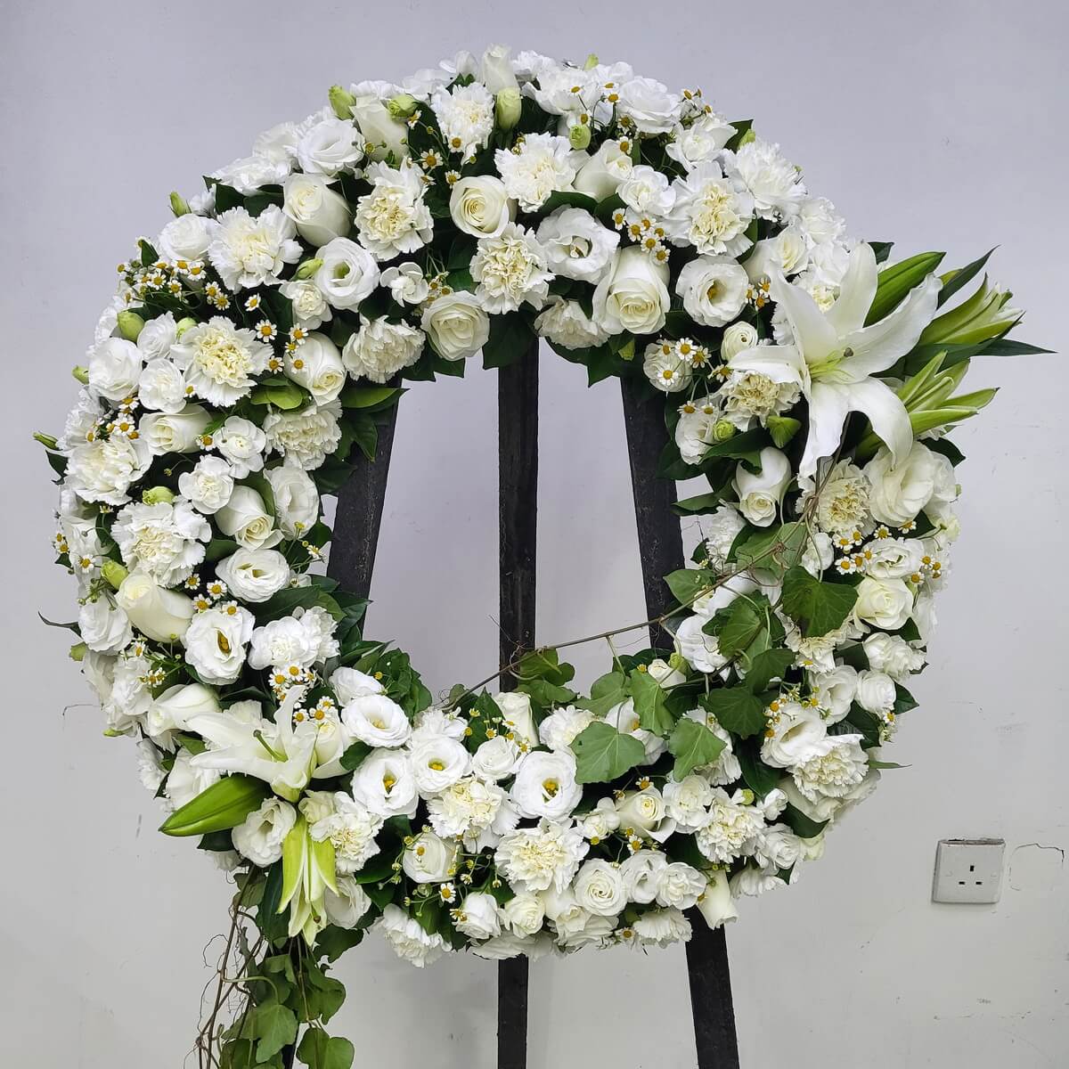 Full floral wreath of white lilies, roses, lisianthus, carnations, yellow-centered chamomile flowers and cascading ivy, displayed on black tripod - 白百合、玫瑰、洋桔梗、康乃馨、黃芯小白菊及垂藤裝飾圓形花圈連黑色三腳支架