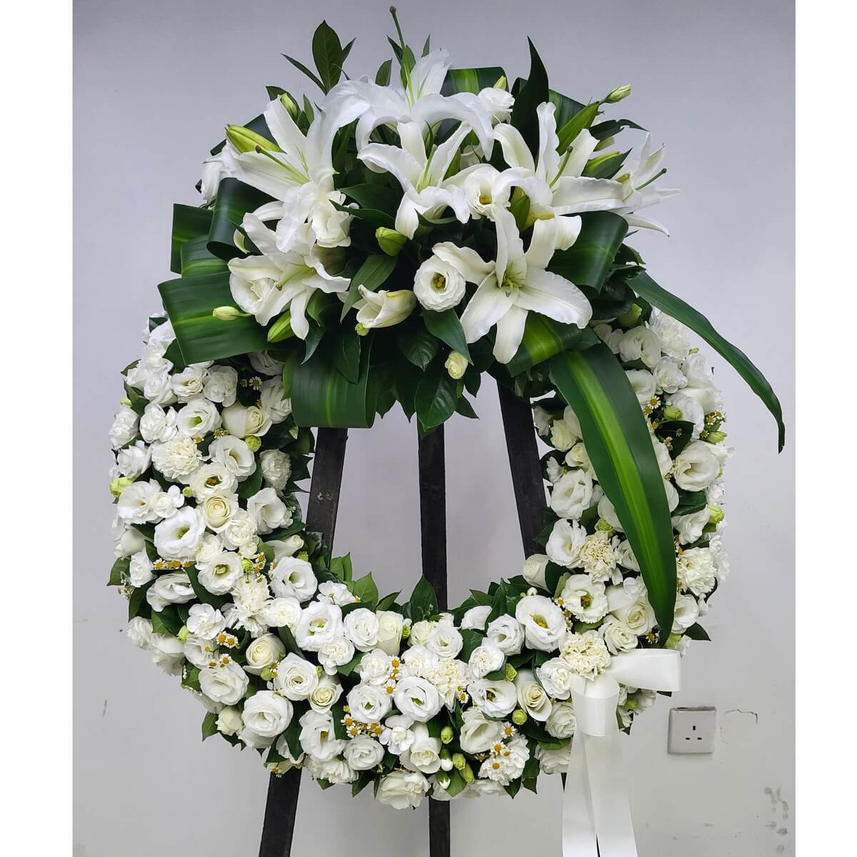 Full wreath with white lilies, roses, lisianthus, carnations, yellow-centered chamomile, structured tropical foliage and a white ribbon, on black stand - 白百合、玫瑰、洋桔梗、康乃馨、黃芯小雛菊及熱帶綠葉配白絲帶圓形花圈連黑色支架展示