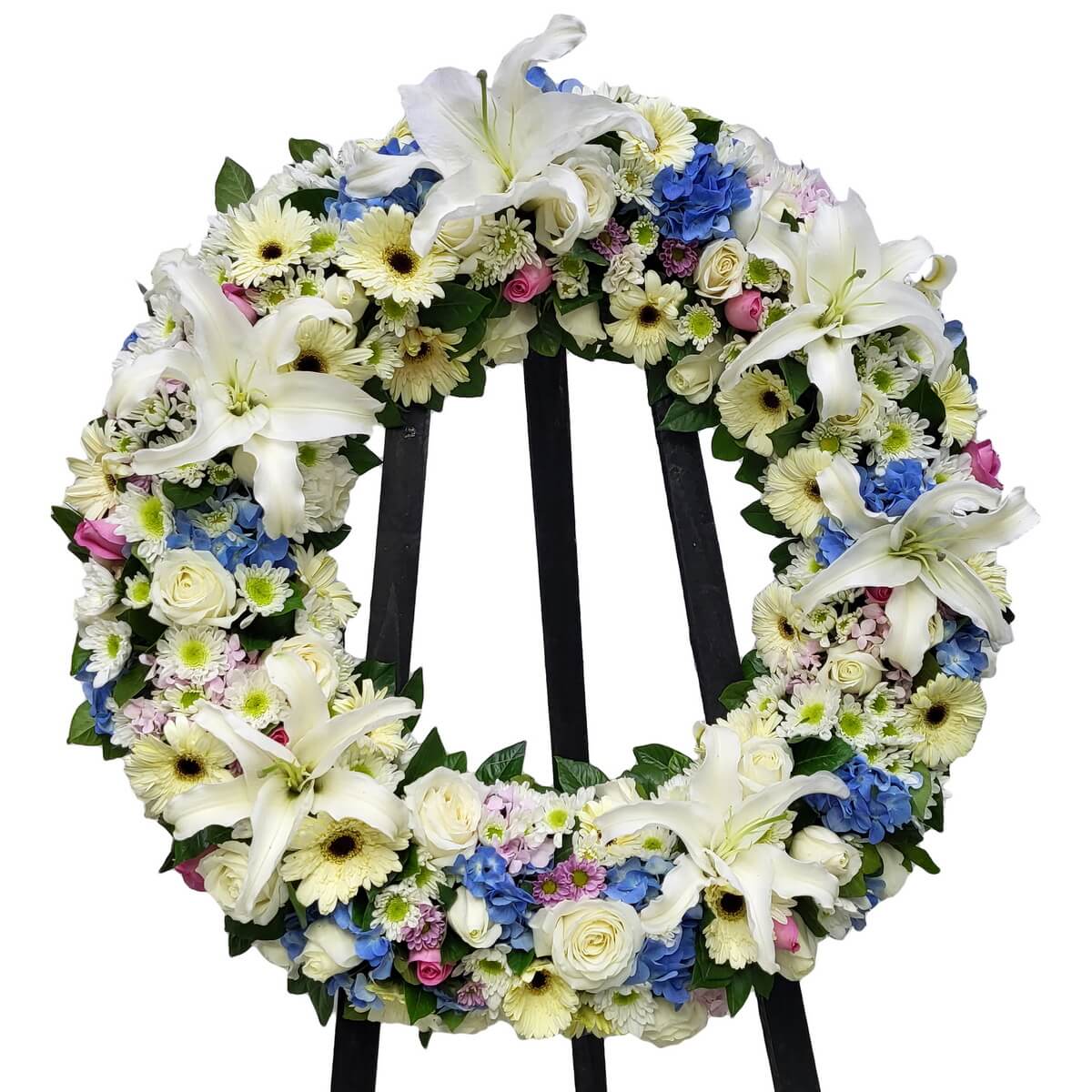 Round wreath featuring white lilies, ivory gerberas, white roses, blue hydrangeas, and pink spray roses. 圓形花圈以白百合、奶油太陽菊、白玫瑰、藍繡球及粉紅玫瑰花束組成，設於黑色支架上。