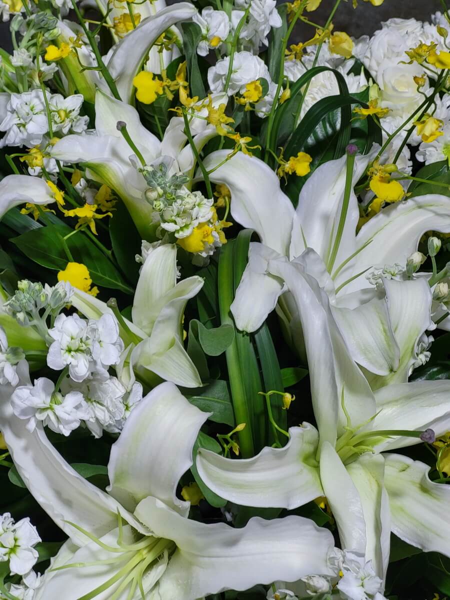 Full view of a mixed floral arrangement featuring white lilies, white stock, and yellow dancing-lady orchids. 花藝作品展示白百合、白宿根花與黃跳舞蘭，搭配深綠葉材，層次分明。