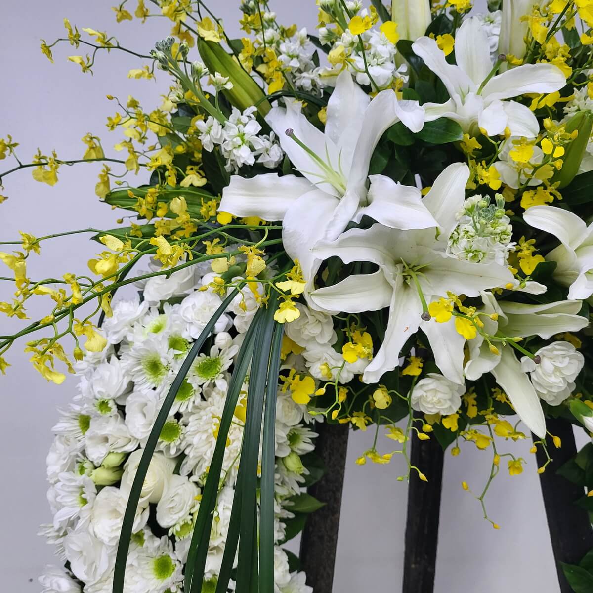 Close-up showing white oriental lilies and yellow dancing orchids arranged with white roses, button chrysanthemums, small stock flowers, and linear greenery 白色東方百合、黃色跳舞蘭配白玫瑰、乒乓菊、小型宿根花與葉材線條的立體特寫