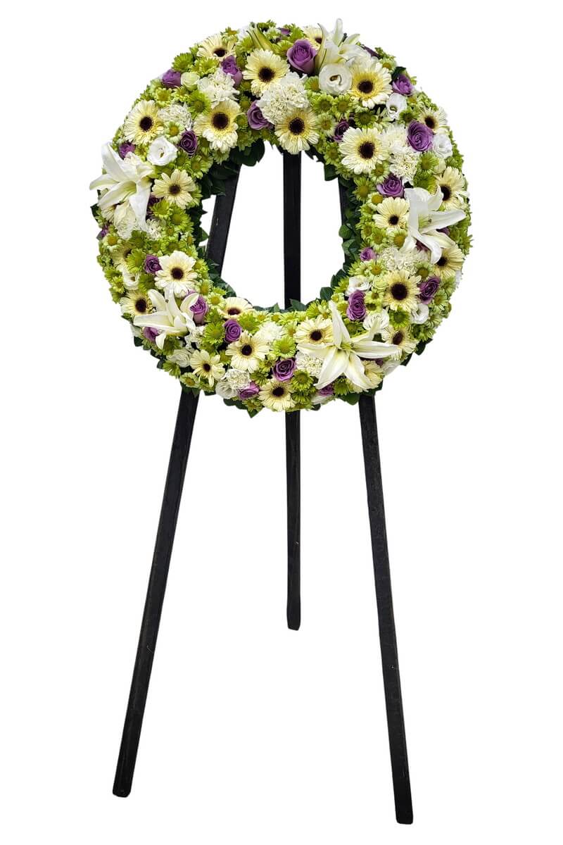 Full floral wreath arranged in a circular form with white gerbera daisies, white lilies, green button chrysanthemums, and purple roses, featuring dense layering and balanced color contrast 白色非洲菊、白色百合、綠色菊花及紫色玫瑰圓形花圈