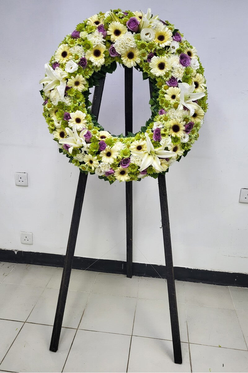 Full circular floral wreath displayed on a stand, composed of white gerbera daisies, white lilies, white lisianthus, green button chrysanthemums, and purple roses with dense floral coverage 白色非洲菊、百合、洋桔梗、綠色菊花及紫色玫瑰花圈設計
