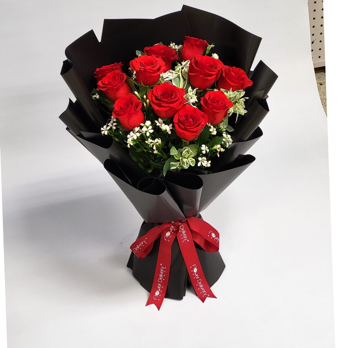 Elegant hand-tied bouquet featuring vibrant red roses, white fillers, and greenery, wrapped in black paper 黑色包裝紙襯托鮮紅玫瑰與白色碎花，搭配綠葉，綁以紅絲帶，展現對比美感與經典浪漫。

