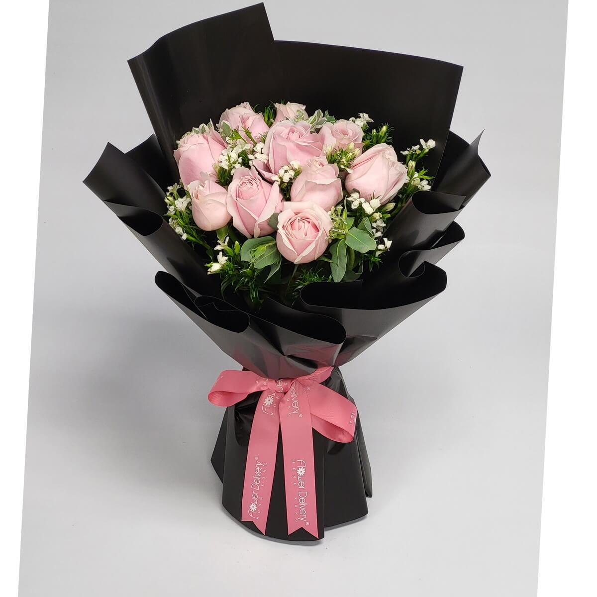 Hand-tied bouquet featuring pale pink roses, white filler flowers, and foliage 淡粉紅玫瑰搭配白色碎花與綠葉，手綁花束以亮黑色包裝紙包裹，粉紅絲帶束口，柔美對比鮮明。