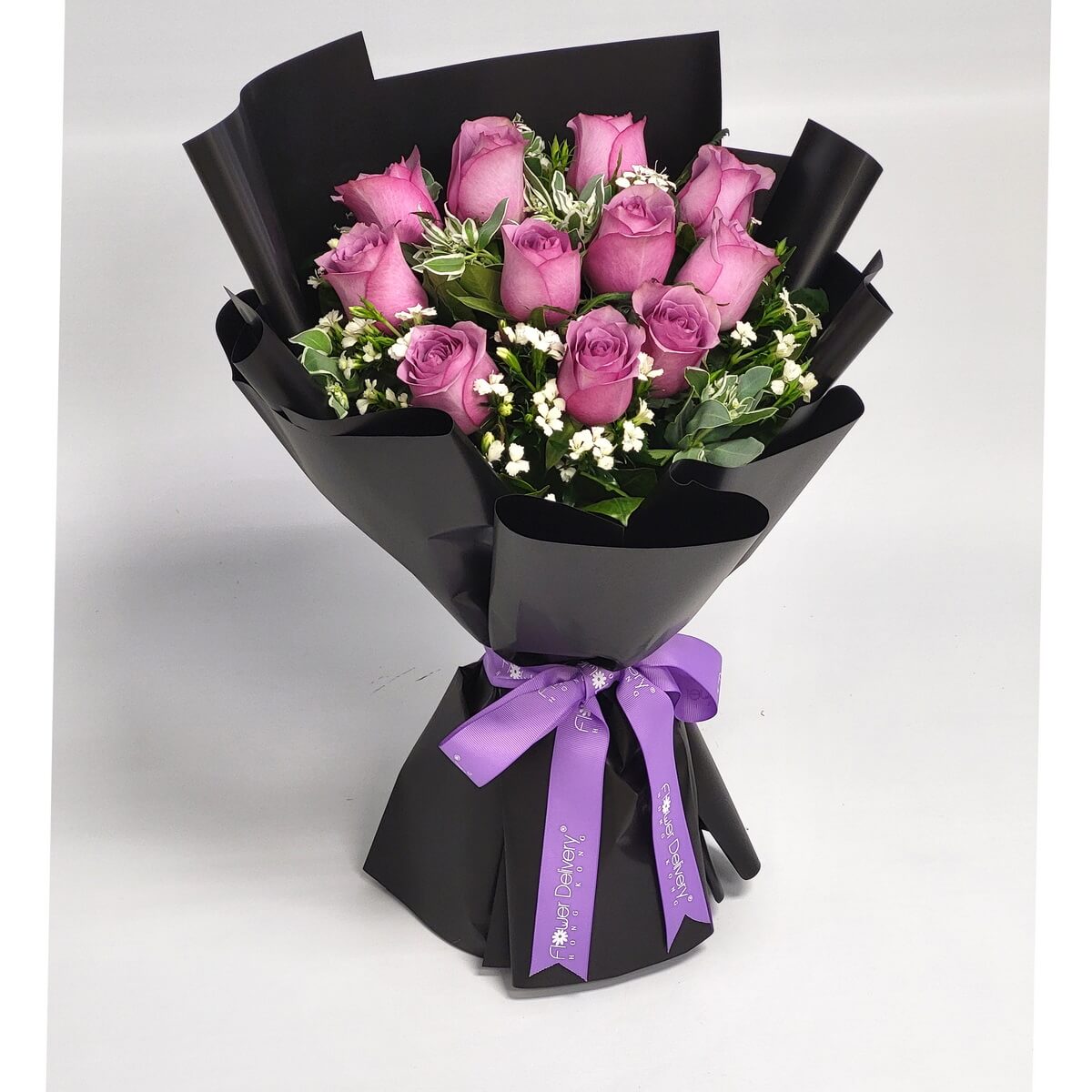Hand-tied bouquet featuring lavender roses, filler flowers, and mixed greenery in black wrap 淺紫玫瑰配白色碎花與葉材，手綁花束設計，包以亮黑色包裝紙及紫色絲帶，造型優雅對比強烈。