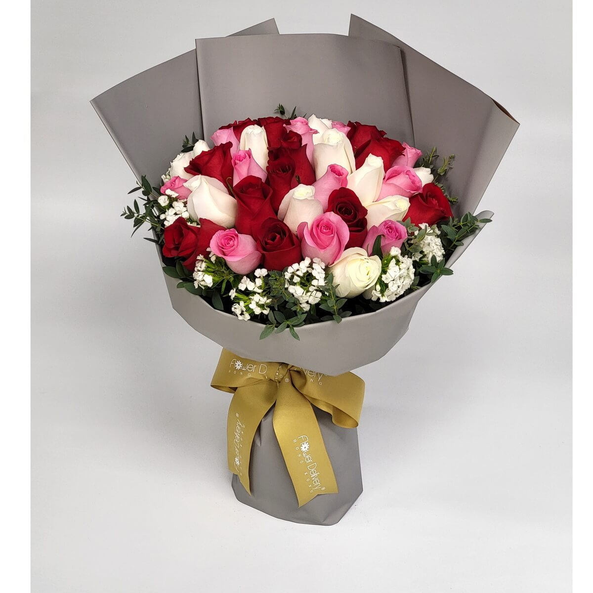 Hand-tied bouquet of red, white, and pink roses with greenery and white filler flowers 紅、白及粉玫瑰環狀排列，搭配白色碎花與綠葉，手綁花束以啞灰色包裝紙包裹，金色絲帶束口，配色優雅。
