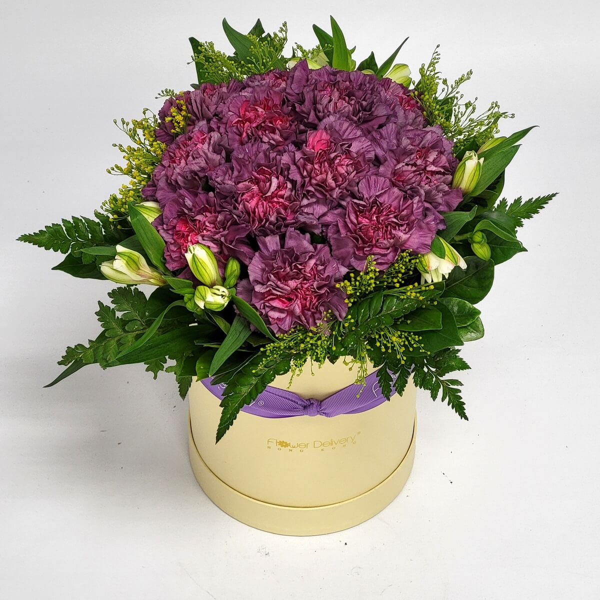 Round box arrangement of purple carnations, white alstroemeria buds, and lush greenery 紫色康乃馨搭配白色六出花花苞與濃綠葉材，設計於米色圓形花盒中，綁以薰衣草紫絲帶。