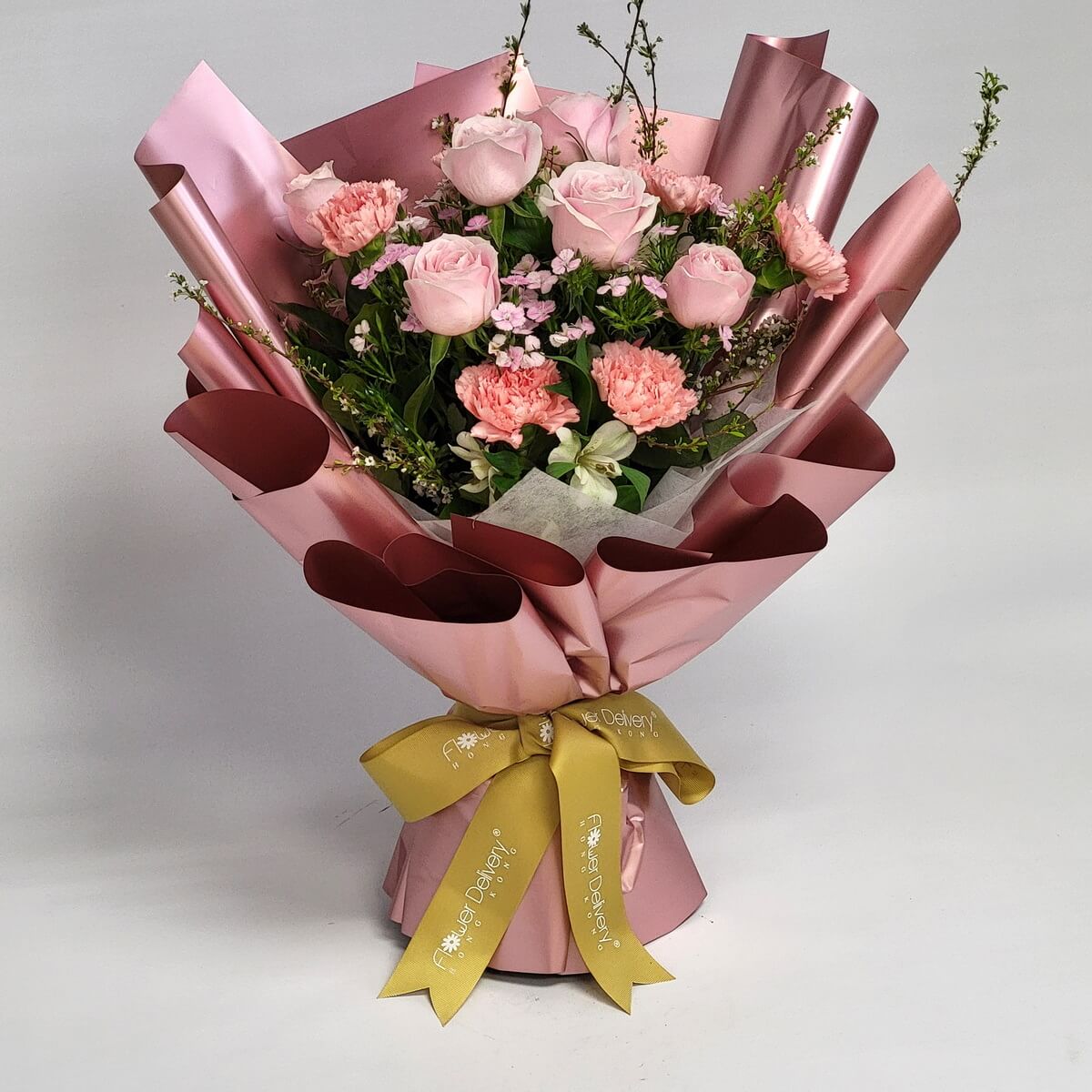 Full blush pink rose and pink carnation bouquet with pink wax flowers, fresh white alstroemeria, and lush seasonal foliage, displayed in layered dusty rose metallic paper with a gold branded ribbon at the base - 淡粉紅玫瑰及粉紅康乃馨全束，配粉紅蠟梅、新鮮白色小百合及豐茂時令葉材，玫瑰金色層疊包裝紙底部繫金色品牌緞帶