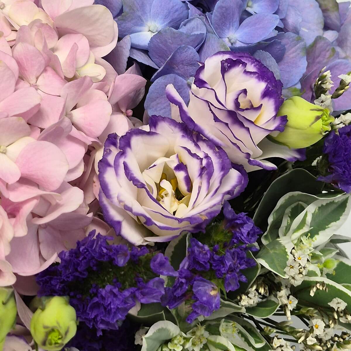 Close-up view of Mother's Day flower arrangement featuring blush pink and blue hydrangeas with white and purple lisianthus, deep purple statice, and seasonal foliage in soft pastel tones - 母親節鮮花禮盒特寫，展示粉紅色及藍色繡球花、白色及紫色洋桔梗、深紫色補血草與季節性綠葉的柔和色調組合