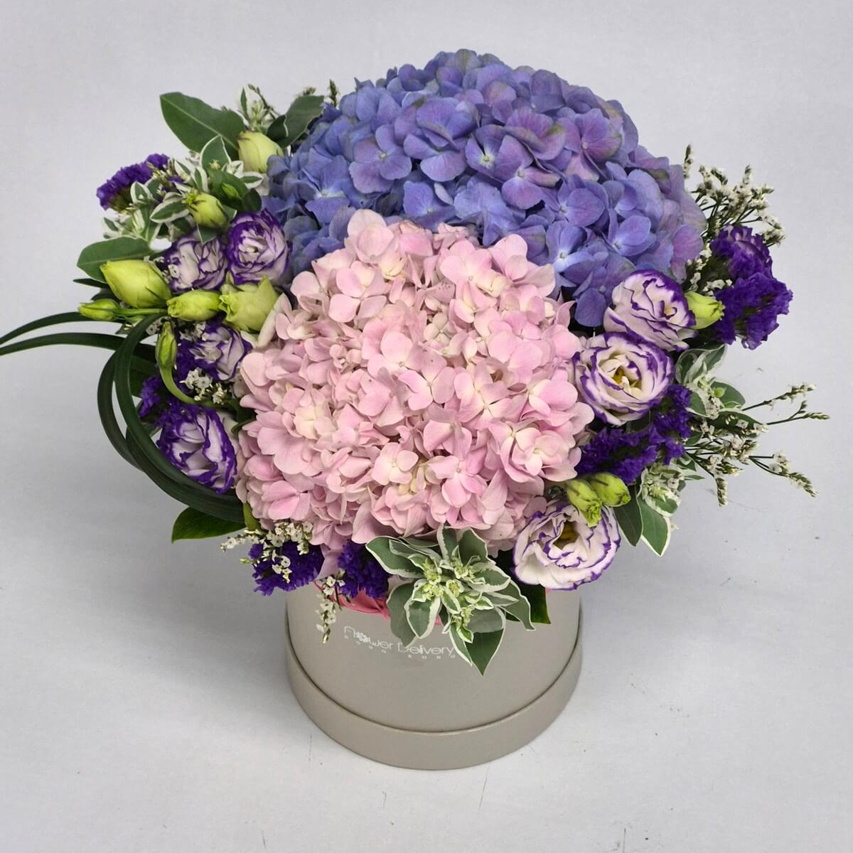 Blue and blush pink hydrangeas with white and purple lisianthus, purple statice, and seasonal foliage arranged in a cream round gift box - 藍色及粉紅色繡球花配白色及紫色洋桔梗、紫色補血草及季節性綠葉，插於奶油色圓形禮盒中的花藝佈置